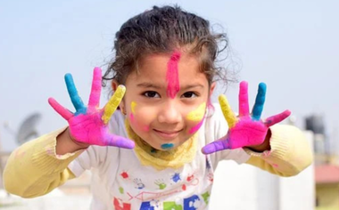 Foto di una bambina sorridente che mostra le mani e il viso sporchi di colori a tempera.