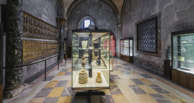 Foto di una sezione del Tesoro di San Marco, con vetrine che espongono alcuni oggetti sacri. Si possono ammirare vasi in vetro colorato, reliquiari in oro e argento, e altri artefatti di valore storico e artistico.