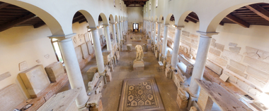 Interno del Museo Nazionale Archeologico Concordiese, con le sue ampie navate sostenute da colonne e i reperti archeologici esposti. Al centro, un mosaico pavimentale e diverse sculture romane.