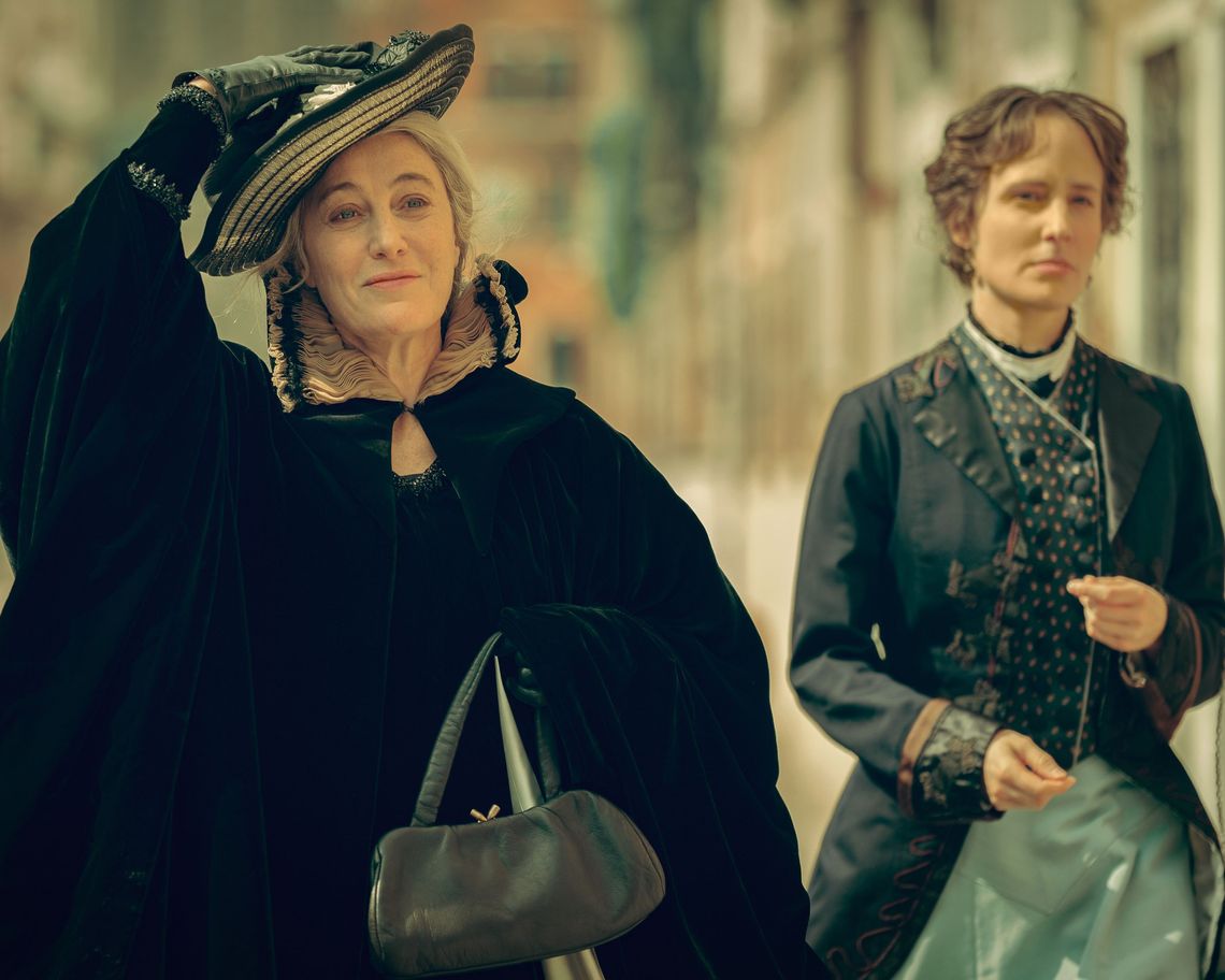 Riprese dalla protagonista che cammina lungo una fondamenta in abito nero tenedo con una mano il cappellino in testa