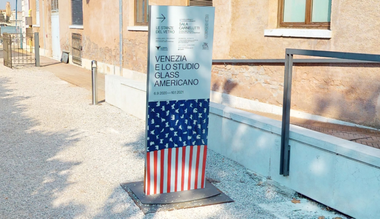 Cartello indicatore esterno della mostra "Venezia e lo Studio Glass Americano", con una freccia che indica la direzione e una grafica a strisce rosse e bianche.