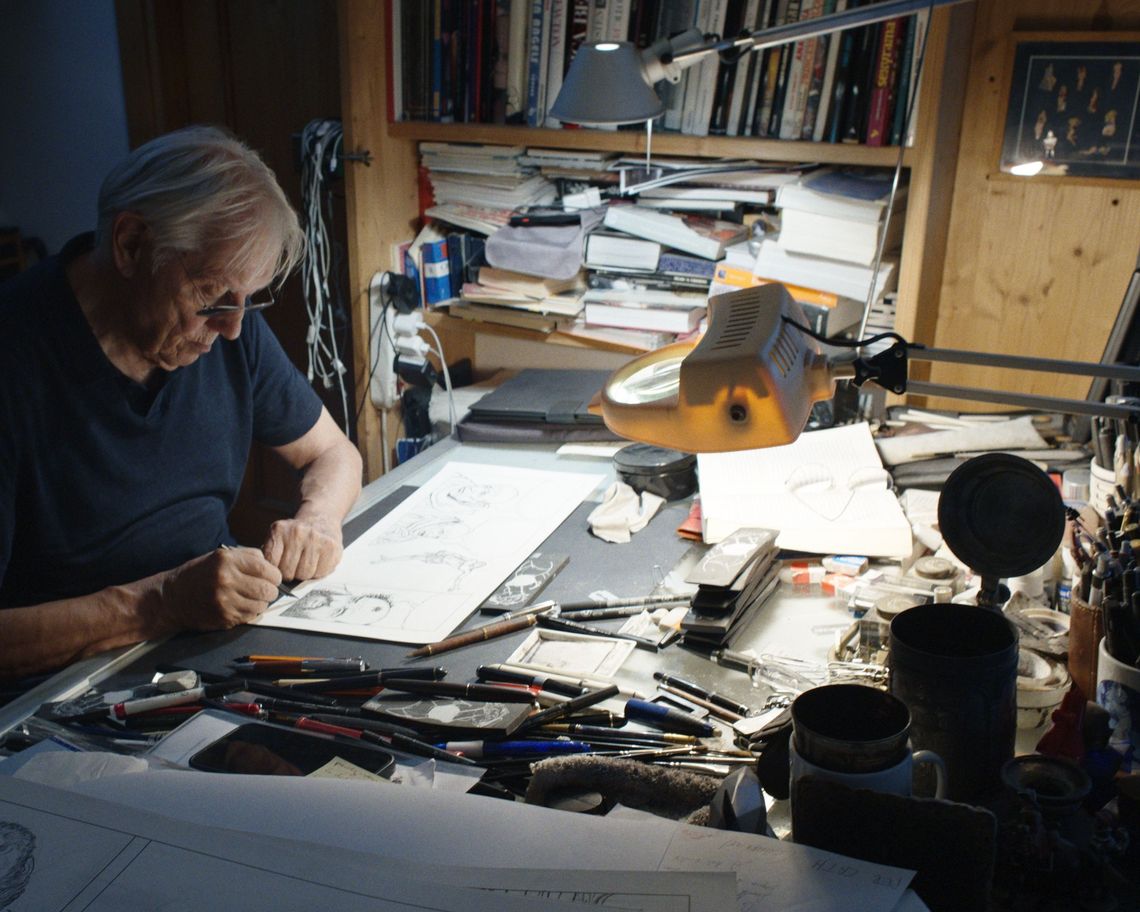 Foto a Milo Manara, artista protagonista del film, piegato sulla scrivania del proprio studio, intento a lavorare. La luce è soffusa, l’ambiente oscurato, mentre l’artista è piegato su bozzetti e fogli sparsi, attorniato dai propri strumenti del lavoro.