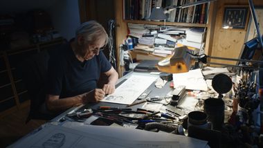 Foto a Milo Manara, artista protagonista del film, piegato sulla scrivania del proprio studio, intento a lavorare. La luce è soffusa, l’ambiente oscurato, mentre l’artista è piegato su bozzetti e fogli sparsi, attorniato dai propri strumenti del lavoro.