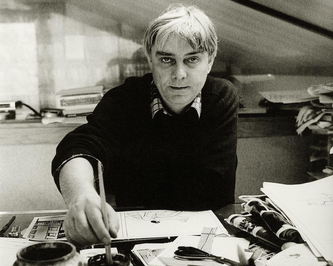 Foto in bianco e nero frontale di Milo Manara alcuni anni fa, intento a lavorare su un bozzetto, con un pennello nella mano destra. La scrivania di fronte a lui è piena di strumenti da pittura e fogli.