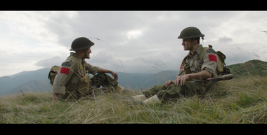 Due soldati stanno seduti uno di fronte all’altro, in una vallata, con le gambe rilassate. Portano con sé le loro armi, indossano la divisa tipica della Seconda Guerra Mondiale color oliva, ma sono intenti in una conversazione rilassata. I due sembrano essere buoni amici. Sullo sfondo i monti verdeggianti e delle nubi in cielo.