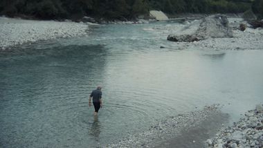 Foto che riprende Marco Paolini dall'alto mentre attraversa a piedi il torrente Cordevole, situato in provincia di Belluno. Lo specchio d'acqua azzurro e grigio, contornato da massi di varia forma e dimensione, lascia intendere l'ambientazione montana. L'uomo, invece, è vestito in modo semplice, con i pantaloni neri arrotolati al ginocchio e le gambe immerse nell'acqua. L'immagine ha toni freddi ed enfatizza la bellezza naturale del territorio montano, oltre alla semplicità di Marco Paolini e del suo amore per esso.