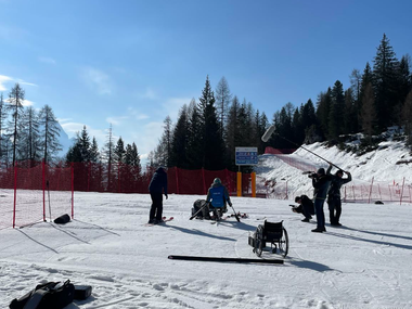 Foto fatta all'esterno, su una grande pista da sci per il film "Over the edge" di Federico Massa e Tommaso Brugin. Il sole è alto in cielo, senza nuvole, così le ombre sul bianco della neve sono definite. Fra le figure presenti sulla scena c'è lo sciatore Renè de Silvestro. Una carrozzina vuota sta in primo piano, mentre sulla destra tre membri della crew fanno le riprese con una cinepresa e microfoni lunghi. Al centro, lo sciatore ha in mano i bastoni che gli serviranno per sciare, mentre un uomo lo aiuto a prepararsi chinato di fronte a lui. Tutti infossano abiti scuri e invernali, mentre lo sciatore indossa abiti da sci colorati di azzurro. La foto li mostra di schiena. Sulla sinistra, un'altra persona indossa degli sci e parla con lo sciatore. Tutt'intorno transenne rosse indicano il limite di sicurezza della pista.