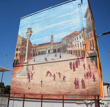 Murale che rappresenta Piazza Vittorio Emanuele di Rovigo, come se fosse una cartolina, dipinta per intero come se fosse vista dall'alto. L'immagine mostra l'intera piazza illuminata da una giornata di sole, con il cielo sgombro, e tutti i simboli architettonici in risalto. Alcune persone sono ritratte in miniatura, in scene di passeggio.