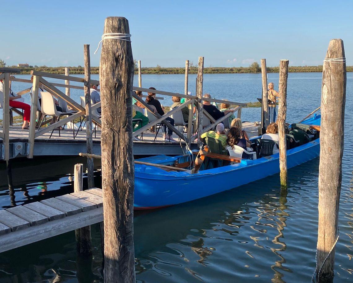una lettrice sul molo legge ad un gruppo di ascoltatori seduti sulle barche