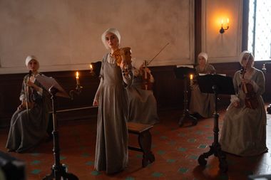 Foto di scena del film Primavera in cui l'attrice Tecla Insolia si trova in mezzo ad una stanza con altre donne. Tutte hanno un violino in mano e sono a una lezione di musica, ma solo lei sta in piedi al centro. La stanza è molto semplice e poco illuminata, le donne portano una specie di abito uniforme, color grigio, e una cuffia bianca, a indicare il loro status umile.