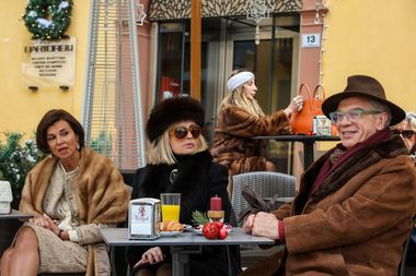 Nella foto si vedono gli attori Isabella Ferrari Marco Marzocca Veronika Logan seduti al tavolino di un bar, all'aperto. I tre indossano abiti invernali piuttosto eleganti: la signora a destra un abito chiaro e uno scialle in pelliccia, la donna al centro un colbacco nero e giacca pesante con pelliccia, l'uomo a destra un cappello stile borsalino e un giaccone dal collo in pelliccia marrone. Dalle decorazioni natalizie sul tavolino e la ghirlanda appesa alla parete dietro di loro si capisce che è il periodo di Natale. Alle loro spalle sta anche un'altra signora, cliente del bar, in abiti invernali e con una fascia bianca scalda orecchie.