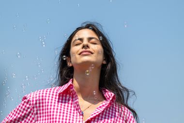 Foto dal basso di Fotinì Peluso, ad occhi chiusi e coi capelli al vento. La giovane donna indossa una camicia a righe bianche e rosa, ha i capelli lunghi castano scuro e leggermente mossi. Il cielo azzurro fa da sfondo alla sua figura e lei ha un'espressione rilassata, godendosi la luce del sole ad occhi chiusi. Delle bolle di sapone le passano davanti.