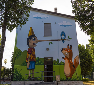Esterno dell'edificio con murales raffigurante Pinocchio con il lungo naso in legno sul quale è appoggiato un uccellino. Pnocchio sta chiaccerando con una volpe.