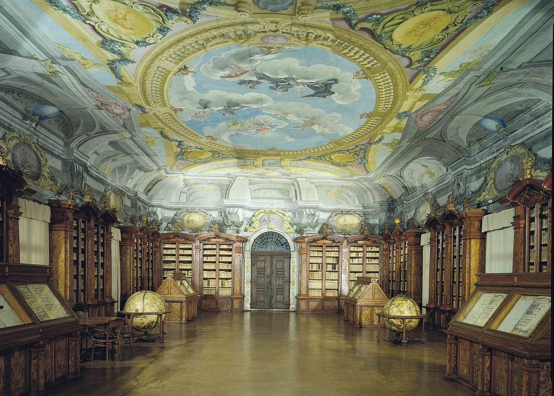 Interno di una delle sale principali della biblioteca con scaffalattura lignea e affresco sul soffitto. Sulle pareti laterali leggii e  mappamondi