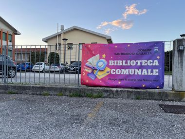 Esterno della Biblioteca Comunale  con ingresso chiuso da un cancello sul quale è appeso un manifesto colorato viola e fucsia con libri e la scritta Biblioteca comunale