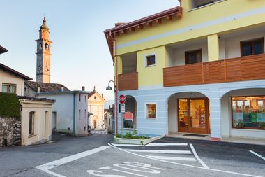 Estreno della biblioteca in edificio moderno a due piani. Il primo paino è bianco con portici e vedrate della biblioteca. Sullo sfondo dell'immagine la chiesa e il campanile.