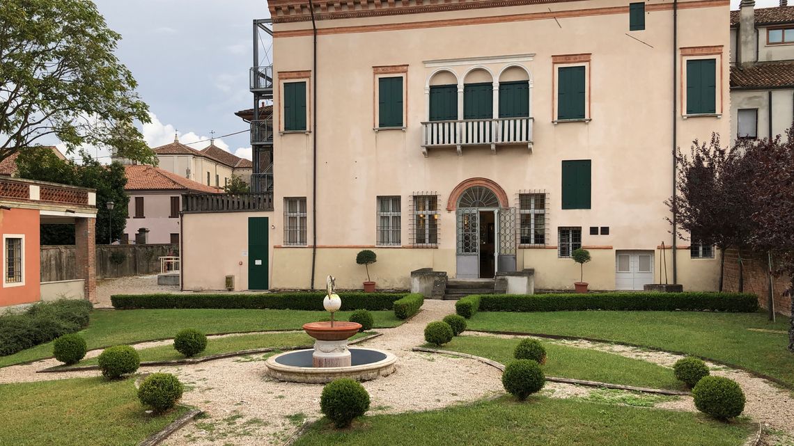 Veduta del giardino e del palazzetto a due piani che ospita la biblioteca. Giardino con fontana centrale e vialetti. Edificio a due piani con balconi verdi e finestre a trifora al secondo piano