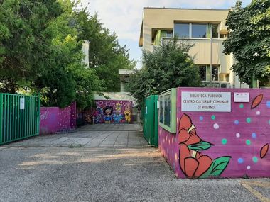 Nell'immagine si vede l'ingresso alla sede della Biblioteca pubblica di Rubano sita in un edificio della seconda metà del Novecento i cui muri di cinta sono decorati con ringhiere e murales dai colori accesi.