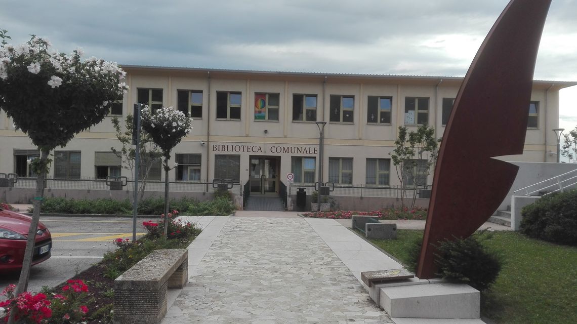 Edificio moderno a due piani preceduto da un vialetto pedonale in pietra, aiuole fiorite con una panchina in pietra, sulla destra una grande scultura in metallo a forma di piuma