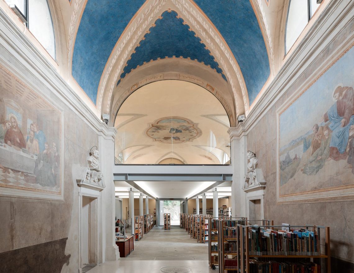 Ambiente interno di una storica ex chiesa caratterizzata da alte pareti decorate, grandi affreschi alle pareti e soffitto a volta con sezioni dipinte di blu. Lungo il corridoio centrale scaffali pieni di libri e colonne strutturali bianche